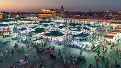 Ein großer Marktplatz bei Nacht, viele überdachte Stände reihen sich aneinander, im Hintergrund breitet sich die Kulisse von Marrakesch aus.
